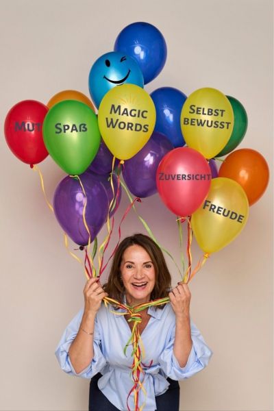 Tanja Funk hält lachend bunte Ballons mit Begriffen wie Mut, Spaß, Freude und Selbstbewusstsein 